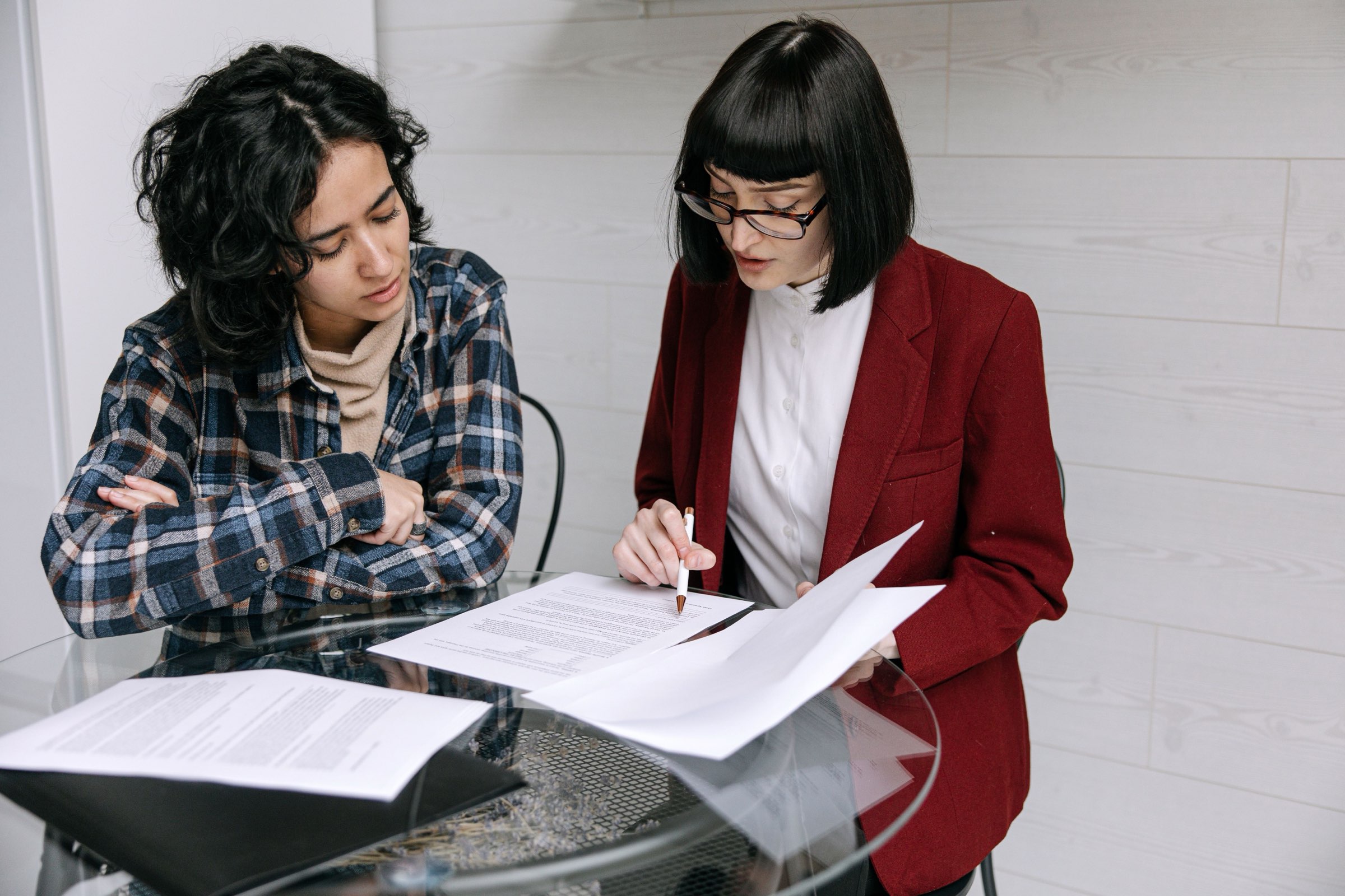 Two people at a glass table reviewing printed contracts, one signing with a pen while the other looks on